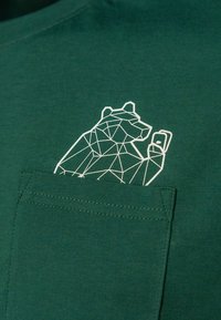 Dunkelgrünes T-Shirt aus Baumwolle mit einem weißen geometrischen Bären-Design über einer Brusttasche. Schlichte und zeitgemäße Textur.