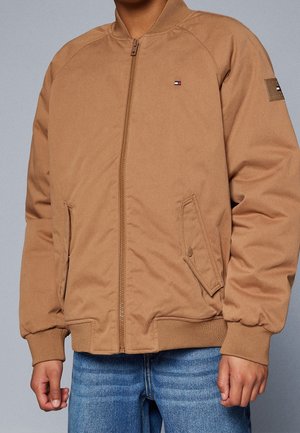 Osoba ubrana w beżową kurtkę typu bomber z przednim zamkiem i ściągaczami przy mankietach, w połączeniu z niebieskimi dżinsami, na jednolitym tle.