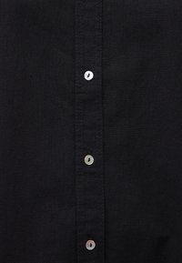 Black shirt with a textured fabric, featuring a row of six pearl buttons down the center and contrasting stitching along the edges.