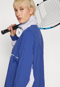 Person mit kurzem blonden Haar trägt eine blau-weiße Jacke und hält einen schwarz-blauen Tennisschläger über der Schulter.
