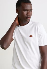 Weißes Baumwoll-T-Shirt mit einem kleinen gestickten Design in Rot und Orange auf der linken Brust. Kurze Ärmel und runder Halsausschnitt.