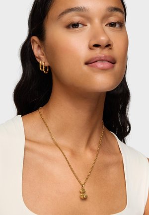 Goldkette mit einem strukturierten Anhänger an einer dünnen Kette, kombiniert mit goldenen Creolen. Das Model trägt ein Oberteil in neutralen Farbtönen und hat einen glatten Teint.