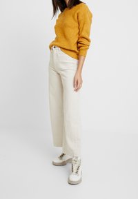 Žlutý pletený svetr s texturovaným vzorem, zkombinovaný s krémovými kalhotami s vysokým pasem a širokými nohavicemi a silnými bílými šněrovacími teniskami.