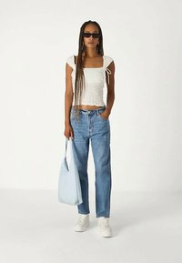 Γυναίκα με μακριά πλεξίματα φορώντας μαύρα γυαλιά ηλίου, λευκό μπλουζάκι με πτυχώσεις, μπλε τζιν, λευκά αθλητικά παπούτσια, κρατώντας μια ανοιχτό μπλε τσάντα.