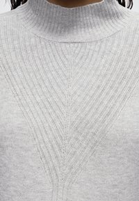 Gros plan d'un pull en maille côtelée gris clair avec un col montant et un motif en chevrons au niveau de la poitrine.