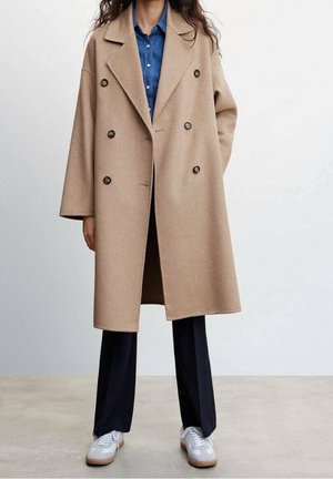 Abrigo beige de doble botonadura con grandes botones negros, solapas anchas y hombros caídos, superpuesto sobre una camisa de mezclilla azul y pantalones azul marino.