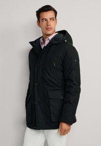 Muškarac u crnoj zimskoj jakni s kapuljačom preko prugaste majice, stoji naslonjen na jednostavnu svijetlo sivu pozadinu, gledajući u stranu.