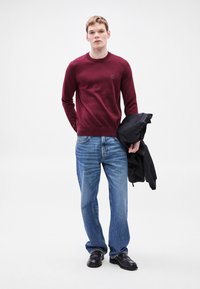 Κόκκινο πουλόβερ με λαιμόκοψη, μπλε τζιν με ίσια μπατζάκια και μαύρα παπούτσια. Το μοντέλο κρατάει μια μαύρη ζακέτα, με λείο ύφασμα και απλό σχέδιο.