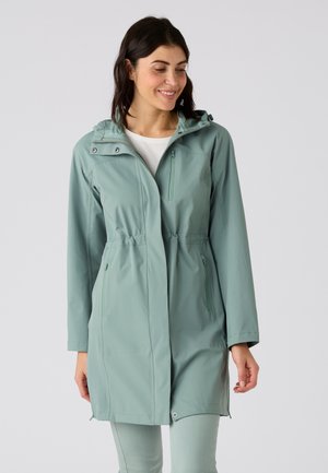 Mujer sonriendo con un chubasquero verde claro con capucha, bolsillos con cremallera y camisa blanca, de pie frente a un fondo claro y simple.