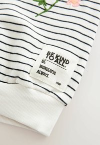 Tissu blanc avec fines rayures noires, fleur rose brodée et un patch blanc portant l'inscription "Be Kind To All. Be Wonderful Always."