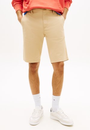 Hombre con pantalones cortos beige hasta la rodilla, zapatillas blancas, calcetines blancos hasta el tobillo y sudadera naranja con las manos en los bolsillos.