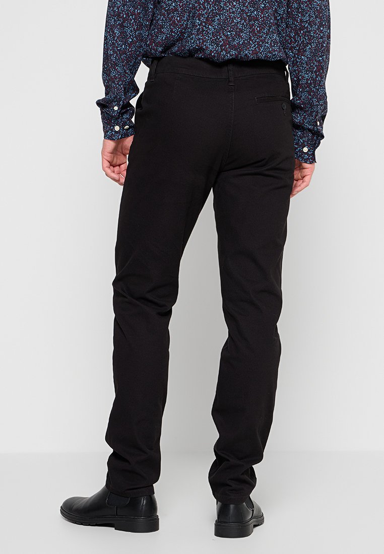 Mann steht mit dem Rücken zur Kamera und trägt schwarze Slim-Fit-Hosen, schwarze Schnürschuhe und ein blaues, gemustertes Langarmhemd.