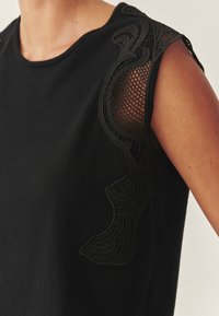 Fekete felső, kerek nyakkivágással és bonyolult fekete csipke részletekkel az egyik vállon, hálós mintázattal és sima szövet textúrával.