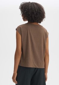 Kvinna med lockigt hår, iklädd en kortärmad brun texturerad topp och svarta byxor, sedd bakifrån mot en enkel vit bakgrund.