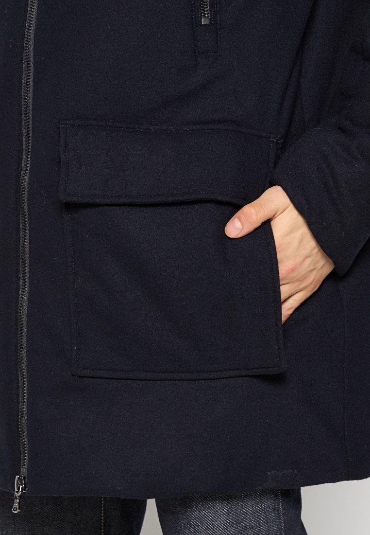 Marineblaue Fleecejacke mit einer großen Fronttasche, Reißverschluss und weicher Textur; gestylt mit einer Hand in der Tasche und dunklen Jeans.