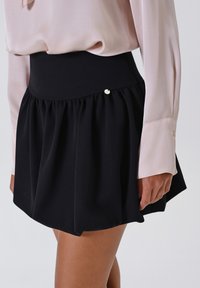 Donna che indossa una gonna nera plissettata a vita alta abbinata a una blusa rosa chiaro a maniche lunghe con ampi polsini.