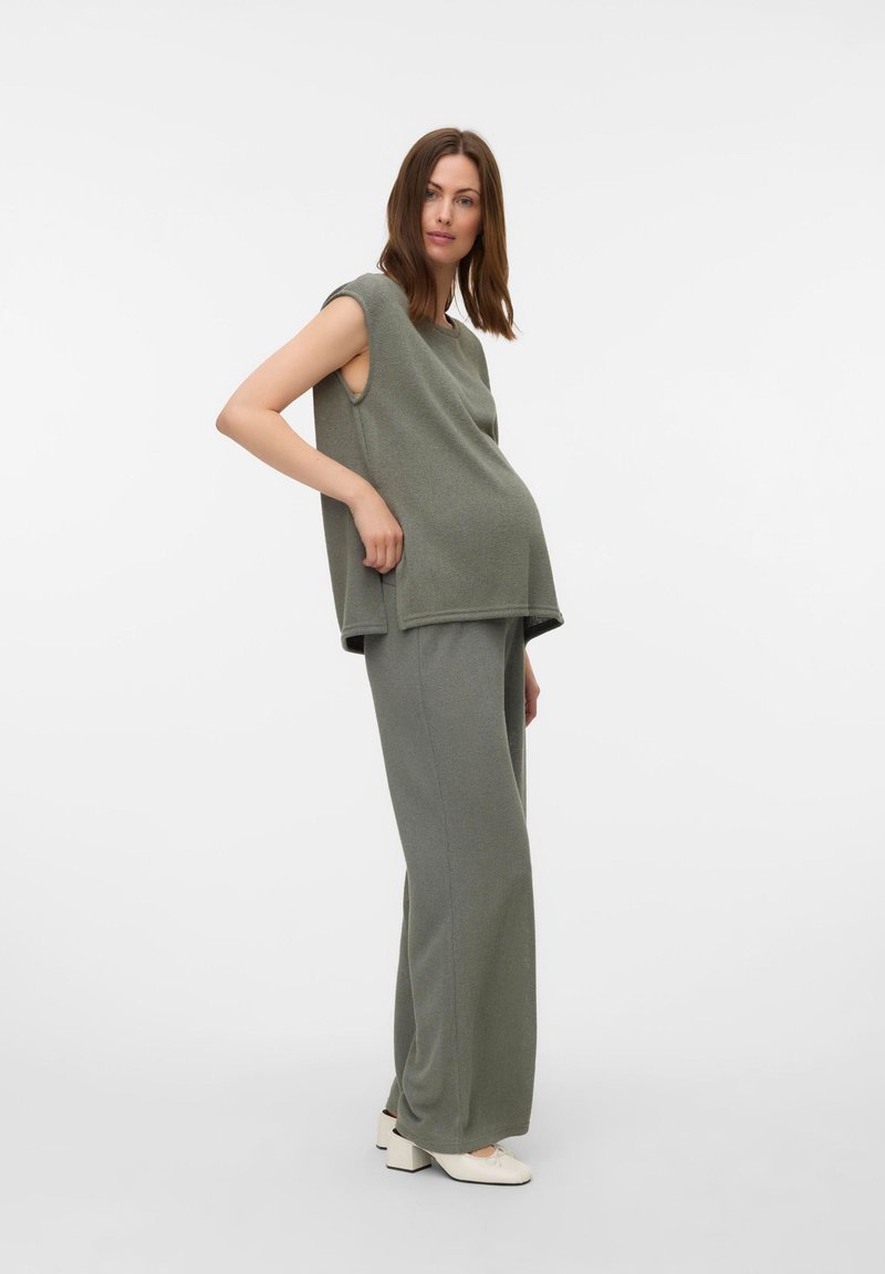 Femme enceinte portant un top ample sans manches et un pantalon ample dans des tons verts atténués avec des chaussures blanches, debout devant un fond blanc uni.