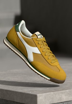 Sneaker giallo con materiali in mesh e suede. Presenta accenti bianchi, un tallone verde e una suola in gomma marrone testurizzata. Design classico.