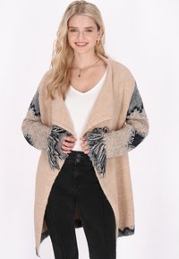 Beige stickad cardigan med grå mönstrade ärmar och fransdetaljer. Bärs över en vit topp och svarta byxor. Har en öppen frontdesign.