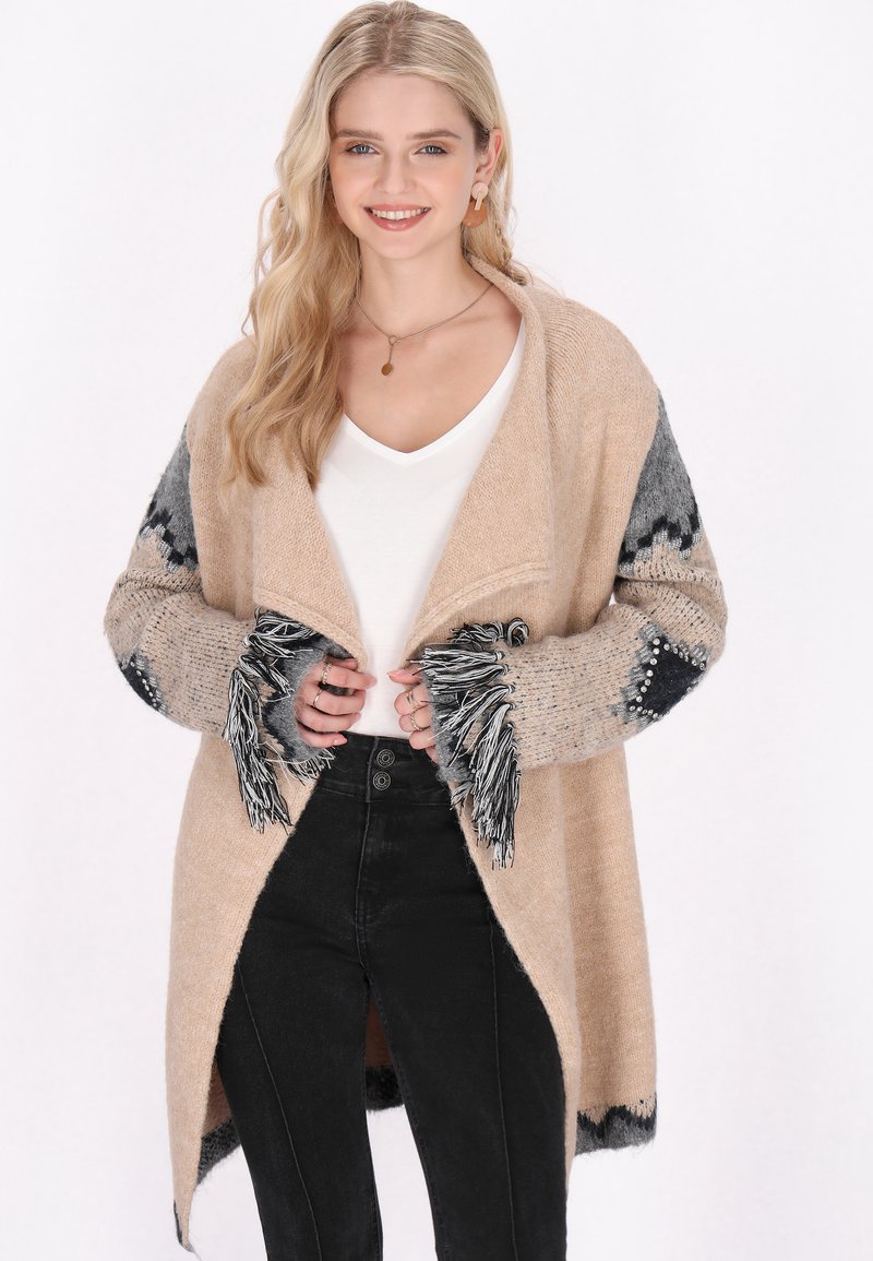 Beige stickad cardigan med grå mönstrade ärmar och fransdetaljer. Bärs över en vit topp och svarta byxor. Har en öppen frontdesign.