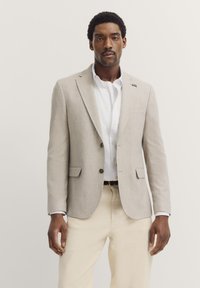 Helles graues Blazer mit zwei Knöpfen, Reverskragen und Seitentaschen, kombiniert mit einem weißen Hemd und beigen Hosen. Glatte Textur.