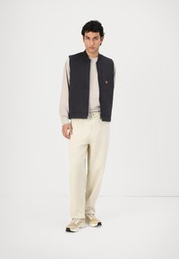 Navy sleeveless vest with a zip closure and side pockets, worn over a beige long-sleeve shirt, paired with cream pants and sneakers.