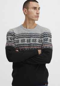 Strickpullover mit einem grauen oberen Abschnitt und einem gemusterten Halsbereich in Schwarz, Rot und Blau, der in die einfarbigen schwarzen Ärmel und den unteren Teil übergeht.