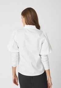 Blusa bianca con maniche a sbuffo e scollatura arricciata. Tessuto liscio e leggero. La vista posteriore mostra una silhouette aderente e la texture delle maniche raccolte.