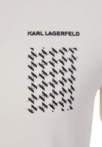 Vit kortärmad skjorta med ett kvadratiskt svart grafiskt mönster med upprepande zigzagformer och texten "KARL LAGERFELD" ovanför.
