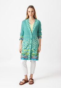 Pitkä turkoosikuvioinen kardigan, jossa on geometrisia muotoja, avoin etuosa ja kolmannes hihat, yhdistetty valkoisiin housuihin ja sandaaleihin.