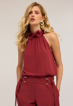 Burgundy sleeveless blouse with a ruffled neckline, paired with matching high-waisted trousers featuring gold hardware accents. Smooth texture.