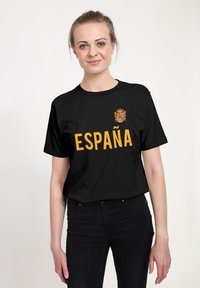 Zwarte katoenen T-shirt met "ESPAÑA" in vetgele tekst en een kleurrijk Spanje-wapen boven de tekst. Korte mouwen en een relaxte pasvorm.
