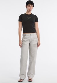 Czarny dopasowany t-shirt z logo "GUESS"; jasnoszare dżinsy proste z kieszeniami z przodu; model nosi czarne japonki i ma krótkie włosy.