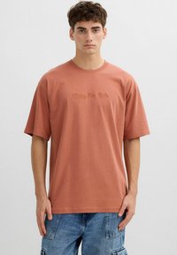 Joven con cabello corto y castaño, vistiendo una camiseta holgada de color óxido con el texto "Disfruta el viaje" y pantalones cortos cargo de mezclilla azul.