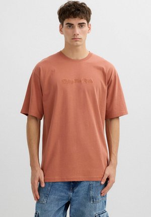 Joven con cabello corto y castaño, vistiendo una camiseta holgada de color óxido con el texto "Disfruta el viaje" y pantalones cortos cargo de mezclilla azul.