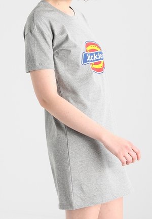 Persoon draagt een grijze korte mouw t-shirtjurk met een cirkelvormig kleurrijk logo en tekst op de borst, staand voor een effen achtergrond.