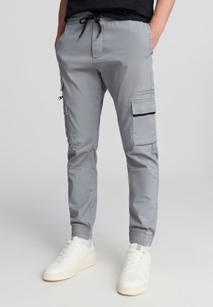 Pantaloni cargo grigi con vita elastica e polsini. Presentano tasche laterali con pattine e una tasca con zip sulla coscia. Tessuto liscio.