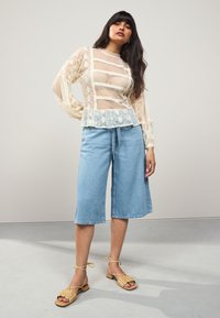 Áttetsző fehér csipkeblúz virágos hímzéssel és fodros szegéllyel, világoskék farmerrövidnadrággal és fonott lapos szandállal párosítva.