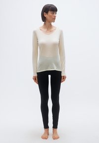 Γυναίκα με κοντά σκούρα μαλλιά στέκεται ξυπόλυτη φορώντας μια κρεμ μπλούζα με μακριά μανίκια και μαύρο κολάν, κοιτάζοντας αριστερά της μπροστά σε απλό φόντο.