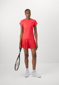 Červené sportovní tričko s krátkými rukávy a plisovanou sukní. Modelka drží tenisovou raketu, má na sobě bílé tenisky a sportovní ponožky.