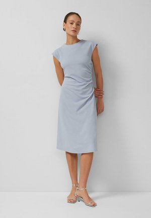 Donna che indossa un vestito azzurro chiaro senza maniche fino al ginocchio con arricciature laterali, orecchini a cerchio argentati e tacchi aperti argentati, in piedi contro un muro bianco.