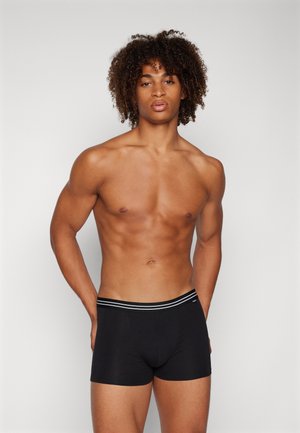 Boxer shorts noirs avec une texture lisse et une ceinture à rayures blanches contrastantes. Le modèle a une silhouette athlétique et des cheveux bouclés.
