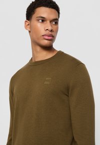 Maglione khaki a maniche lunghe con scollatura rotonda, polsini a costine e un'etichetta quadrata piccola con la scritta "BOSS" nella zona sinistra del petto.