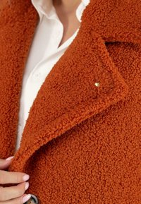 Oranger Texturmantel aus einem fuzzy, leicht strubbeligen Material. Er verfügt über einen Reverskragen und einen dekorativen goldenen Nieten nahe der Revers.