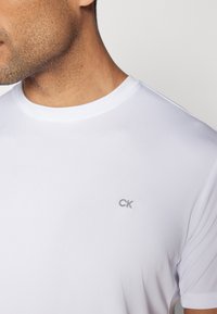 Bela bombažna majica s kratkimi rokavi in okroglim izrezom, ki ima majhen sivi logo "CK" na levi strani prsnega koša.