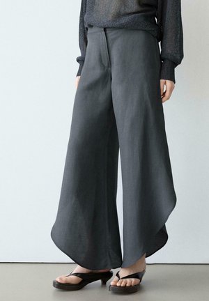 Personne portant un pantalon à jambes larges gris foncé avec des ourlets incurvés et des sandales à talons noires, debout devant un fond clair uni.