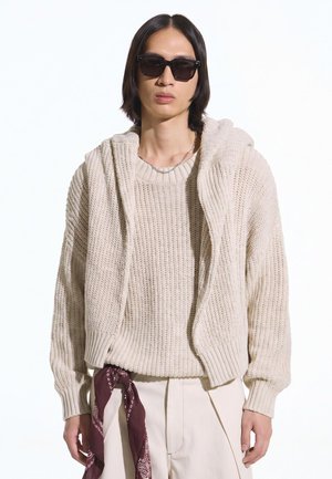 Jong persoon met zwarte zonnebril, beige grofgebreide trui en jas, crèmekleurige broek met een donkerrode bandana die uit de zak hangt.