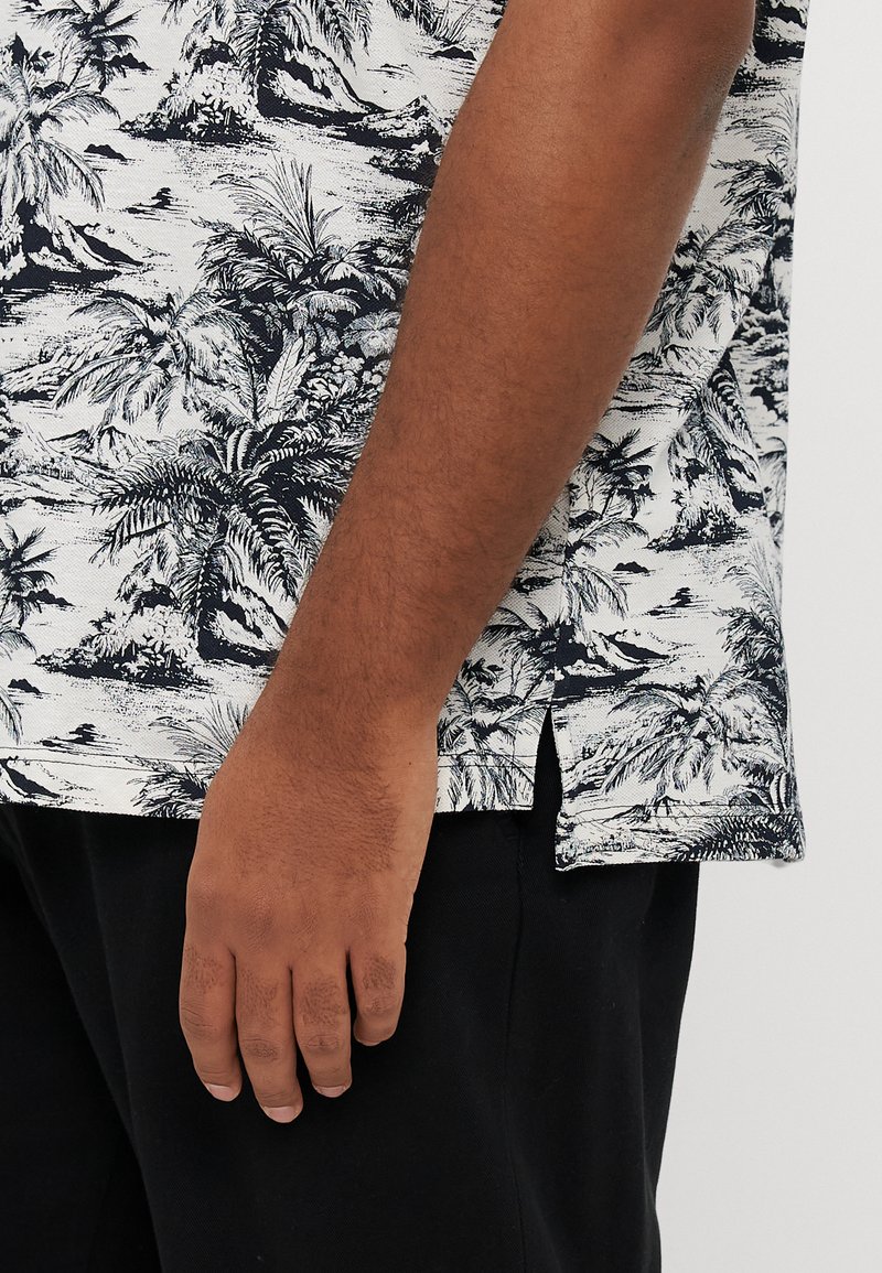 Kurzärmeliges Hemd in Weiß mit schwarzem Palmenprint. Der Stoff ist leicht und der Schnitt entspannt. Seitliche Schlitze am Saum sorgen für Bewegungsfreiheit.