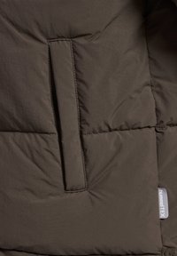 Bruine pufferjack sectie met een rechthoekige zak, gestikte textuur en een merklabel. Materiaal lijkt lichtgewicht en is geïsoleerd.