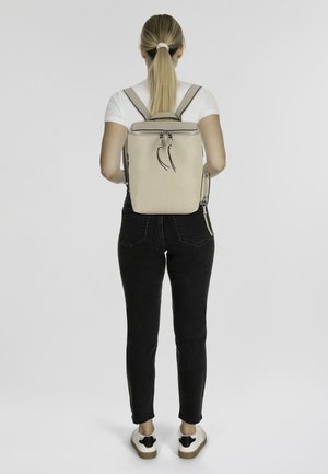 Tansfarbener Leder-Rucksack mit strukturiertem Design, doppelten Reißverschlüssen und verstellbaren Trägern, getragen von einer Person, kombiniert mit eng anliegenden schwarzen Jeans und Sneakers.